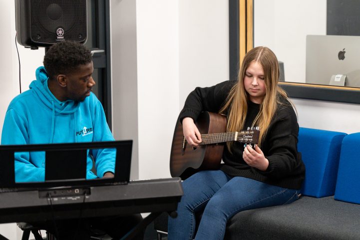Katie is sat in the music room with music youth worker, Clint. Katie is strumming on the guitar and Clint is playing the keyboard
